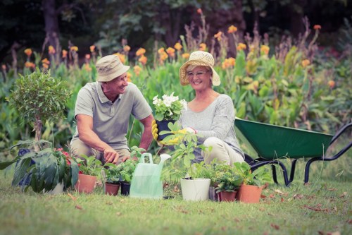 A middle-aged man and woman seated together in a lush garden during daytime, both wearing straw hats and gardening gloves, smiling as they handle blooming plants. The garden features a well-maintained grassy lawn in the foreground, with various potted plants placed on the grass, including flowering and foliage varieties. Behind them, vibrant purple and orange flowers grow densely along a border, with taller green shrubs and trees providing a natural backdrop. To the right, there's a dark green garden bench and a light green watering can among the plants. The scene suggests outdoor gardening activities, highlighting the enjoyment of caring for garden plants in a residential outdoor space, with natural sunlight creating a warm and inviting atmosphere. The environment appears peaceful and well-maintained, reflecting professional gardening and landscaping practices common in Cricklewood and surrounding areas.