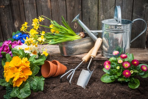 The image shows a composted garden bed with dark, rich soil in the foreground. To the left, there are colorful flowering plants including yellow primroses, purple pansies, and white daffodils, creating a vibrant display. On the right, a potted plant with pink and yellow daisies is visible. Behind the plants, gardening tools such as a hand rake, garden trowel, and fork rest on the edge of a metal container that holds additional gardening implements. An empty terracotta plant pot lies on the soil near the plants. In the background, a weathered wooden fence runs horizontally, providing privacy for the garden space. The scene appears to be set on a cloudy day with soft, diffuse lighting, highlighting the variety of textures and colours typical of a well-maintained outdoor garden area. This setting exemplifies typical gardening and landscaping activities handled by professionals such as Hedge Trimming Cricklewood as part of comprehensive garden maintenance services for residential outdoor spaces in the Cricklewood area, ideally suited to local gardening needs.